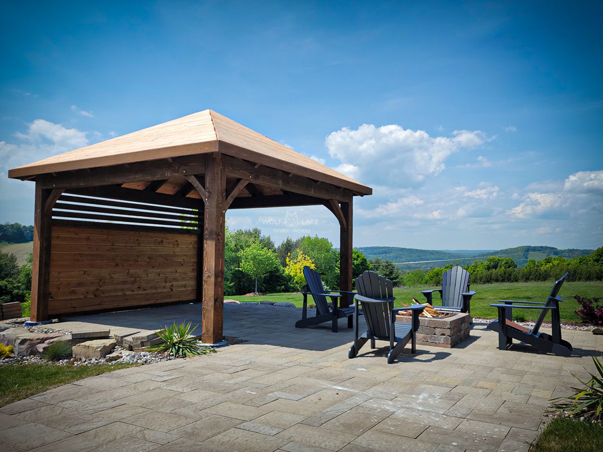 A wooden pavilion with a slatted back wall overlooking rolling hills, surrounded by Adirondack chairs and a fire pit.